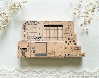 May include: A set of 10 wooden stamps with various designs, including a to-do list, a calendar, a piggy bank, stars, a bird, a sun, a cloud, and a picture frame. The stamps are arranged in a staggered pattern on a white surface.