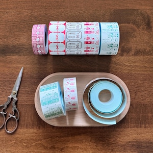 May include: Overhead view of decorative washi tape rolls with diverse designs. The tape features patterns such as coffee tickets, trains, and baseballs. A wooden tray holds more rolls, and a pair of scissors sits on the wooden surface.