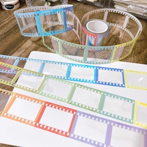 May include: A roll of decorative washi tape and strips of film strip tape. The tape features a clear design with colorful gradients of blue, green, orange, and purple. The film strip tape has the text "ORANGE PORTRA" and numbers.