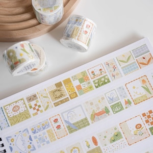 May include: A roll of washi tape with a repeating pattern of colorful squares and floral designs. The tape is on a white surface with other rolls of washi tape.