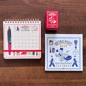 May include: A selection of stationery items with a retro postal theme, including a spiral-bound calendar, a small red box, and a square card. The calendar shows the month of January 2026. The items are arranged on a wooden surface.