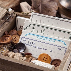 Può includere: Una scatola di legno piena di etichette di carta in stile vintage e bottoni. Le etichette hanno accenti blu e verdi e un testo che dice "HILD", "PHARMACIE FELIX", "No. 1599", "SCEIVED with thanks from" e "195".