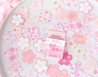 May include: A sheet of pink and white decorative stickers featuring a cherry blossom design. The stickers are arranged in a random pattern on a white background. The text on the stickers reads "Sakura Whisper".