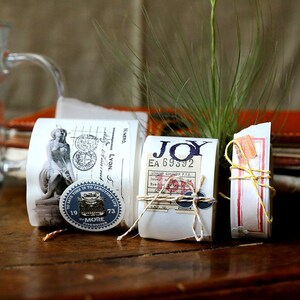 May include: Three rolls of decorative washi tape with different designs. The first roll features a vintage illustration of a woman, the second roll has the word "JOY" printed on it, and the third roll has a red and white striped design. The rolls are wrapped with twine and have small paper tags attached.