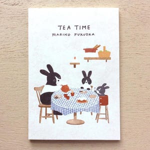 Puede incluir: Una tarjeta blanca con el texto "TEA TIME" y "MARIKO FUKUOKA" en letras negras. Una ilustración de tres conejos negros y blancos sentados a una mesa con un mantel a cuadros, tomando el té y comiendo pastel.