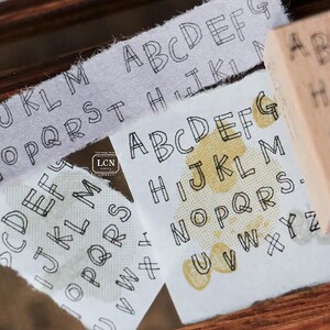 May include: Close-up of printed alphabet letters on fabric strips and a wooden stamp. The letters are in a simple, outlined font. The fabric has a textured appearance, and the stamp is visible in the background.