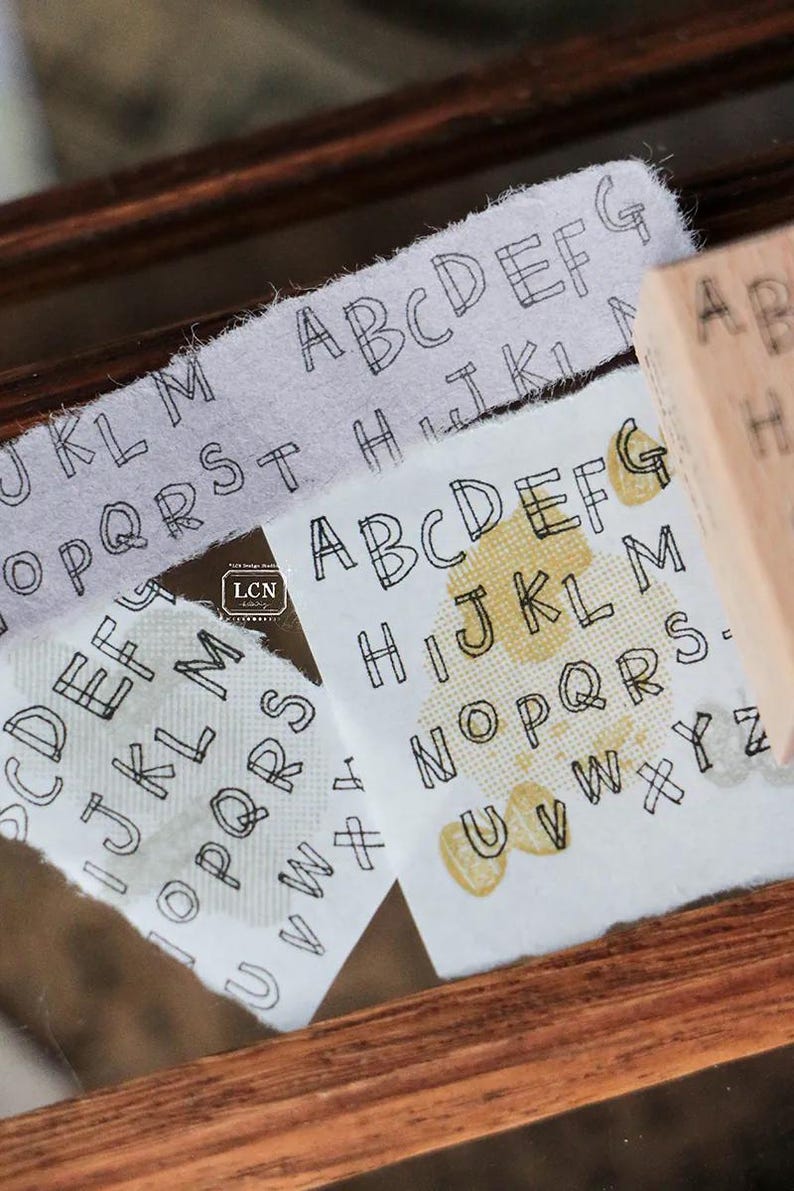 May include: Close-up of printed alphabet letters on fabric strips and a wooden stamp. The letters are in a simple, outlined font. The fabric has a textured appearance, and the stamp is visible in the background.