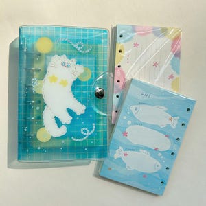May include: A clear blue grid-patterned binder featuring a white cat illustration with yellow star accents, accompanied by two sets of loose-leaf paper inserts, one with pastel clouds and one with fish-shaped diet tracking graphics.