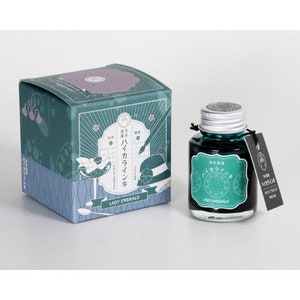 May include: A bottle of Lady Emerald ink, a teal green ink with a green and white label. The label features Japanese text and a stylized image of a hat. The bottle is sitting on a white surface.