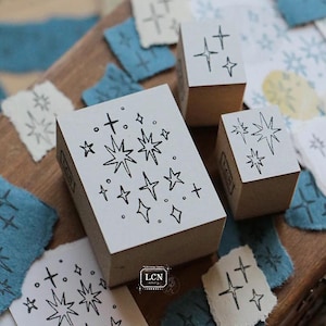 May include: Three wooden rubber stamps featuring star and sparkle designs. The largest rectangular stamp displays a cluster of various star shapes, while two smaller square stamps feature simpler star patterns. The text LCN is visible on the stamps.