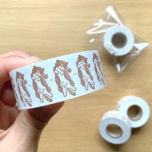 May include: White washi tape roll featuring a repeating pattern of brown cat illustrations. Additional rolls of tape are visible, one in a clear plastic bag and two loose. The tape is designed for crafts, scrapbooking, or decorative purposes.