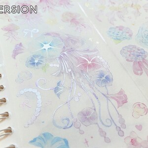 Puede incluir: Una hoja de cinta washi transparente con un acabado hologr&aacute;fico que presenta un patr&oacute;n repetitivo de medusas azul pastel y rosa, flores y remolinos. El texto "WASHI VERSION" est&aacute; impreso en la parte superior de la hoja.