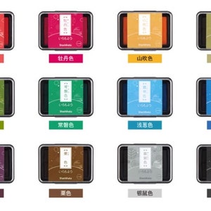 May include: A collection of twelve ink pads in various colors, each in a rectangular black case. The pads feature Japanese text and color names, including red, pink, orange, green, blue, and purple.