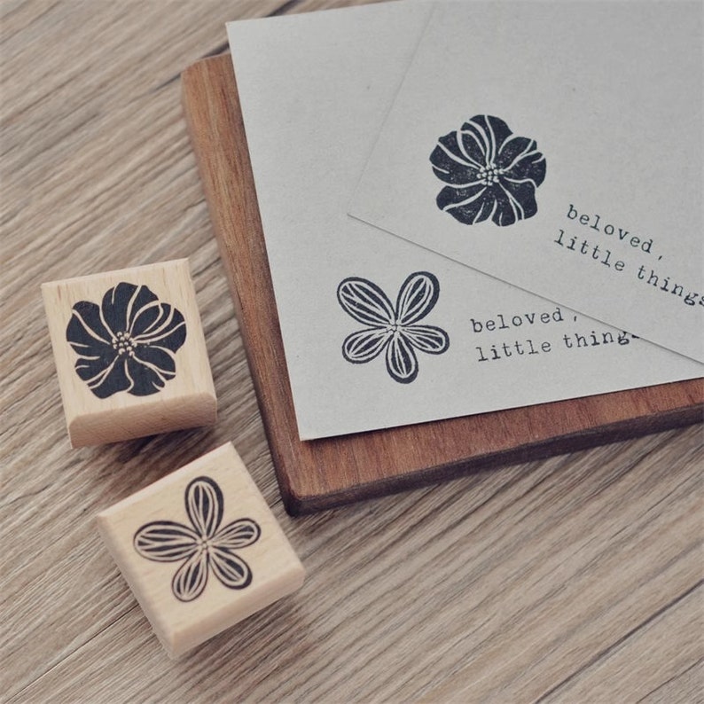 May include: Three wooden stamps with black floral designs. The stamps are on a wooden surface. The text "beloved, little things" is printed on a card.