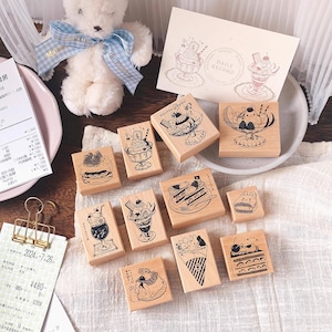 Puede incluir: Una colección de sellos de madera con ilustraciones de postres, incluyendo helados, pasteles y parfaits. Los sellos están dispuestos sobre una superficie blanca, con un oso de peluche y un recibo en el fondo. También hay una tarjeta con el texto "DAILY RECORD".