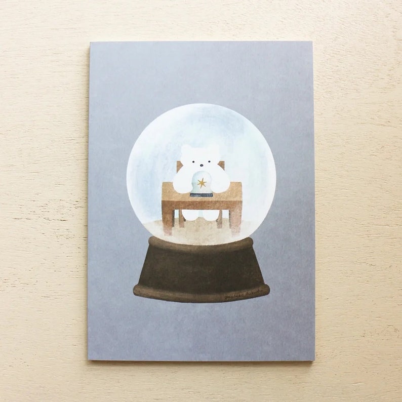 Puede incluir: Un oso polar blanco con un sombrero azul y dorado se sienta en una silla de madera dentro de una bola de nieve de vidrio. La bola de nieve tiene una base marr&oacute;n.