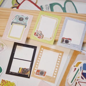 May include: A selection of decorative paper cards featuring camera illustrations. The cards have different designs, including frames and camera graphics in blue, red, and yellow. The cards are arranged on a wooden surface.