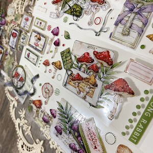 May include: A sheet of clear stickers with various designs, including mushrooms, leaves, and a vintage-style letter. The stickers are printed with a watercolor effect and have a glossy finish. The text "LETTER" and "MUSHROOM" are visible on the stickers.