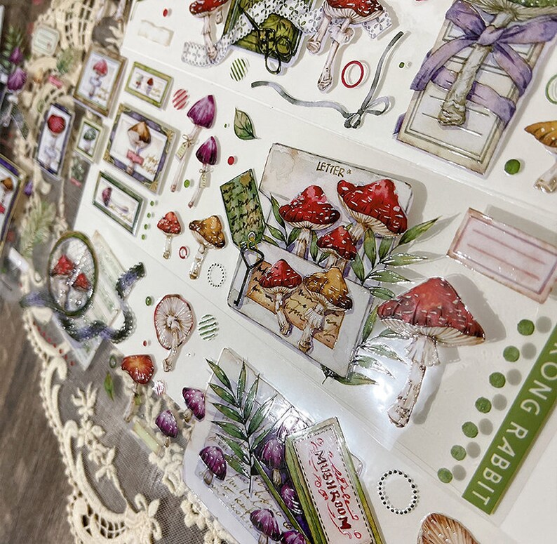 May include: A sheet of clear stickers with various designs, including mushrooms, leaves, and a vintage-style letter. The stickers are printed with a watercolor effect and have a glossy finish. The text "LETTER" and "MUSHROOM" are visible on the stickers.