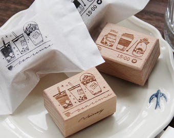 May include: Three wooden stamps with illustrations of different beverages, including coffee, tea, and milk. The stamps are rectangular and have a light brown color. The text "tea time" is engraved on two of the stamps.