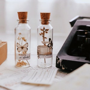 May include: Two clear glass bottles with cork stoppers, each containing dried flowers and a butterfly. One bottle has a butterfly and the text "ce Label". The other bottle has a butterfly and the text "Alexandra Machen". A vintage typewriter is in the background.