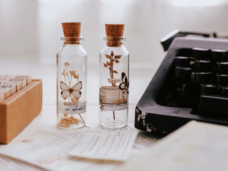 May include: Two clear glass bottles with cork stoppers, each containing dried flowers and a butterfly. One bottle has a butterfly and the text "ce Label". The other bottle has a butterfly and the text "Alexandra Machen". A vintage typewriter is in the background.