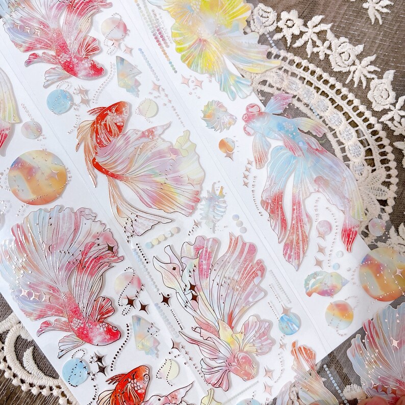 May include: A sheet of colorful, iridescent stickers featuring a variety of fish designs. The stickers are perfect for adding a touch of whimsy to journals, planners, and other craft projects.