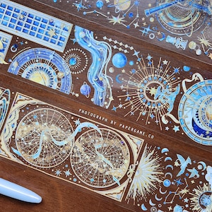 May include: Decorative washi tape rolls featuring celestial and astronomical designs in shades of blue and gold. The tape includes illustrations of planets, stars, constellations, and astronomical instruments. The text "PHOTOGRAPH BY PAPERGAME.CO" is visible.