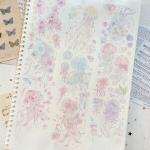 Puede incluir: Una hoja de papel de pegatina transparente con ilustraciones de acuarela coloridas de medusas y flores. Las medusas son transl&uacute;cidas y tienen tent&aacute;culos largos y flotantes. Las flores son en tonos de rosa, morado y azul.