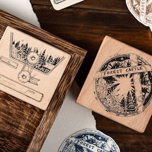 Puede incluir: Dos sellos de madera con diseños de tinta negra. Un sello presenta una escena de bosque con una cinta y el texto "To, From, Date". El otro sello tiene un diseño circular con el texto "FOREST CASTLE" y una escena de bosque.