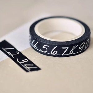 May include: Black washi tape with white numbers 4 through 9 printed on it. The tape is rolled up and sitting on a beige surface.