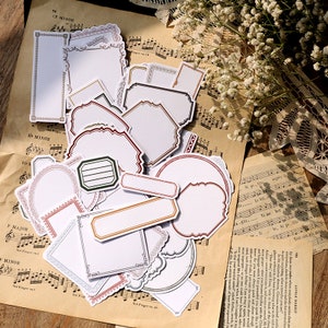 May include: A collection of vintage-style paper stickers in various shapes, including rectangles, ovals, and irregular forms. The stickers feature decorative borders in colors like red, gold, and green. They are arranged on a background of aged sheet music.