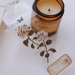 May include: A clear tape roll featuring a floral design with white and light purple flowers and green leaves. A brown glass jar candle with the text "Oakwood & Tobacco" is visible. A small label with the word "Registration" is also present.