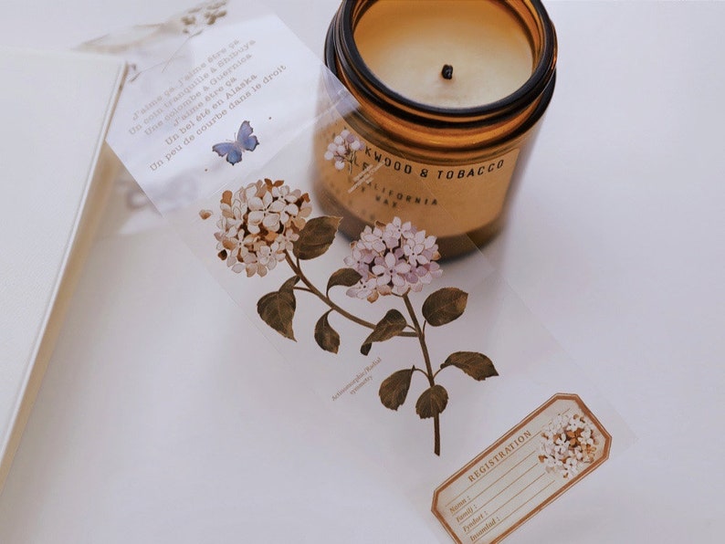 May include: A clear tape roll featuring a floral design with white and light purple flowers and green leaves. A brown glass jar candle with the text "Oakwood & Tobacco" is visible. A small label with the word "Registration" is also present.