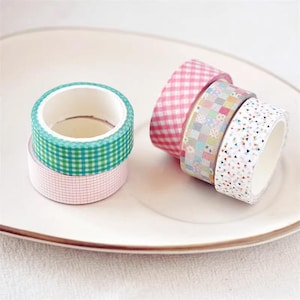 Può includere: Assortimento di nastri washi a motivi colorati, tra cui gingham rosa, patchwork e bianco con piccoli triangoli. I nastri sono arrotolati e mostrati su un piatto bianco.