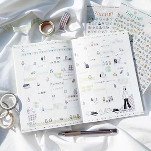 May include: An open planner with a monthly calendar view. The planner is decorated with colorful washi tape and stickers featuring small animals and houses. The planner is open to the month of February.