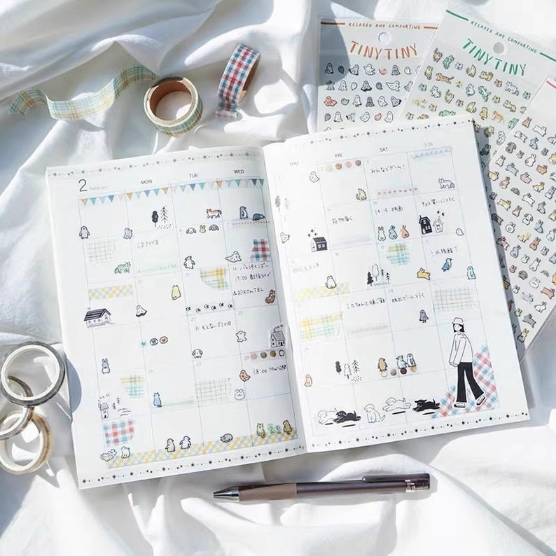May include: An open planner with a monthly calendar view. The planner is decorated with colorful washi tape and stickers featuring small animals and houses. The planner is open to the month of February.