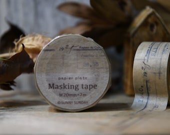 May include: Close-up of two rolls of masking tape with vintage-style labels. The tape is beige with printed text, including "Masking tape" and measurements of W20mm x 7m. The rolls are partially unrolled, revealing the tape.