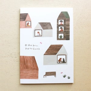 Puede incluir: Una libreta blanca con una ilustraci&oacute;n colorida de casas con personas mirando por las ventanas. El texto "O sha beri notebook" est&aacute; impreso en la portada de la libreta.