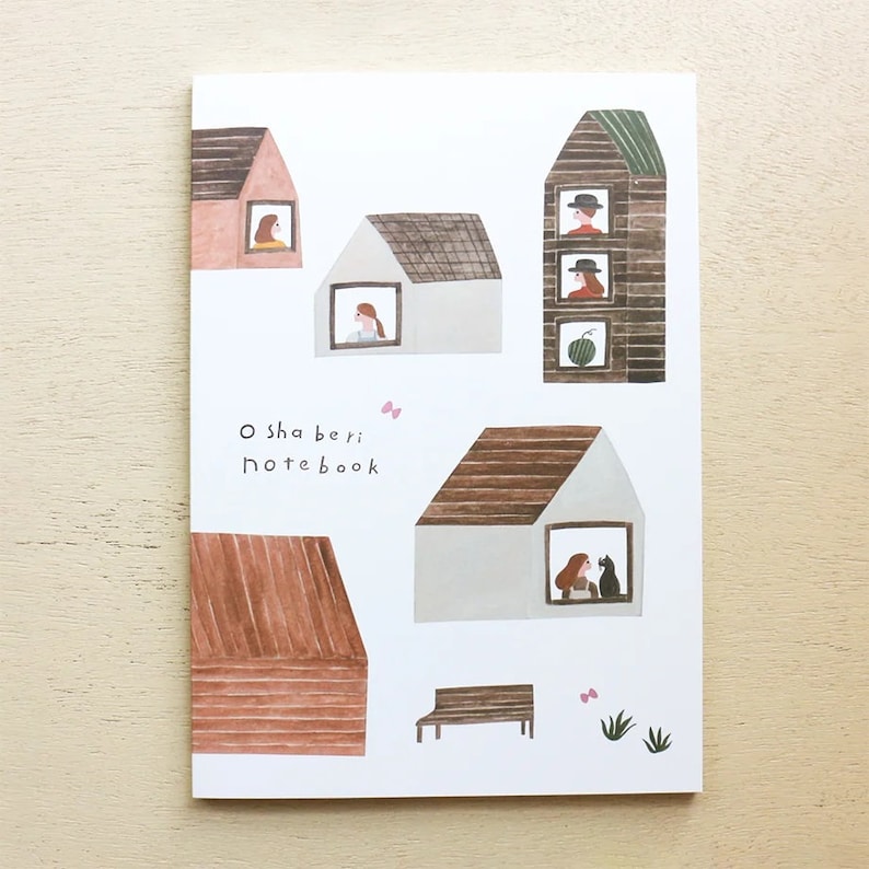 Puede incluir: Una libreta blanca con una ilustraci&oacute;n colorida de casas con personas mirando por las ventanas. El texto "O sha beri notebook" est&aacute; impreso en la portada de la libreta.