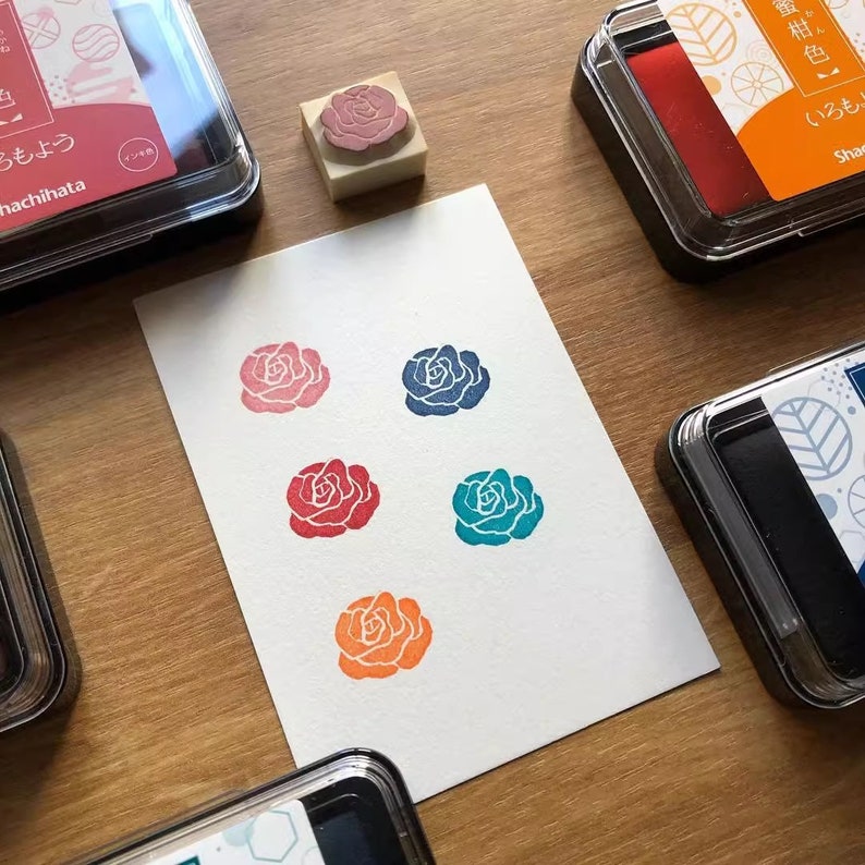 May include: A rose-shaped stamp and ink pads in various colors. The stamp has a detailed rose design, and the ink pads are in red, blue, teal, orange, and pink. The stamped roses are on a white paper, showcasing the ink colors.