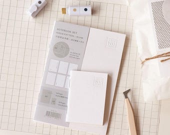 May include: A stationery set featuring a notebook, notepad, and two white and gold decorative clips. The notebook set includes a design pattern and blank pages. The items are arranged on a grid-lined surface.