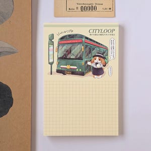 Pode incluir: Um bloco de notas retangular com um padrão de grade cor de creme. A parte superior apresenta um autocarro verde com a etiqueta CITYLOOP, uma paragem de autocarro e um coelho de desenho animado com chapéu e vestido. Texto japonês e Let's Go.