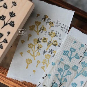 May include: Wooden floral stamp next to stamped paper with the alphabet and numbers. The paper has a torn edge and is stamped with yellow and blue floral designs. The stamp has a floral design.