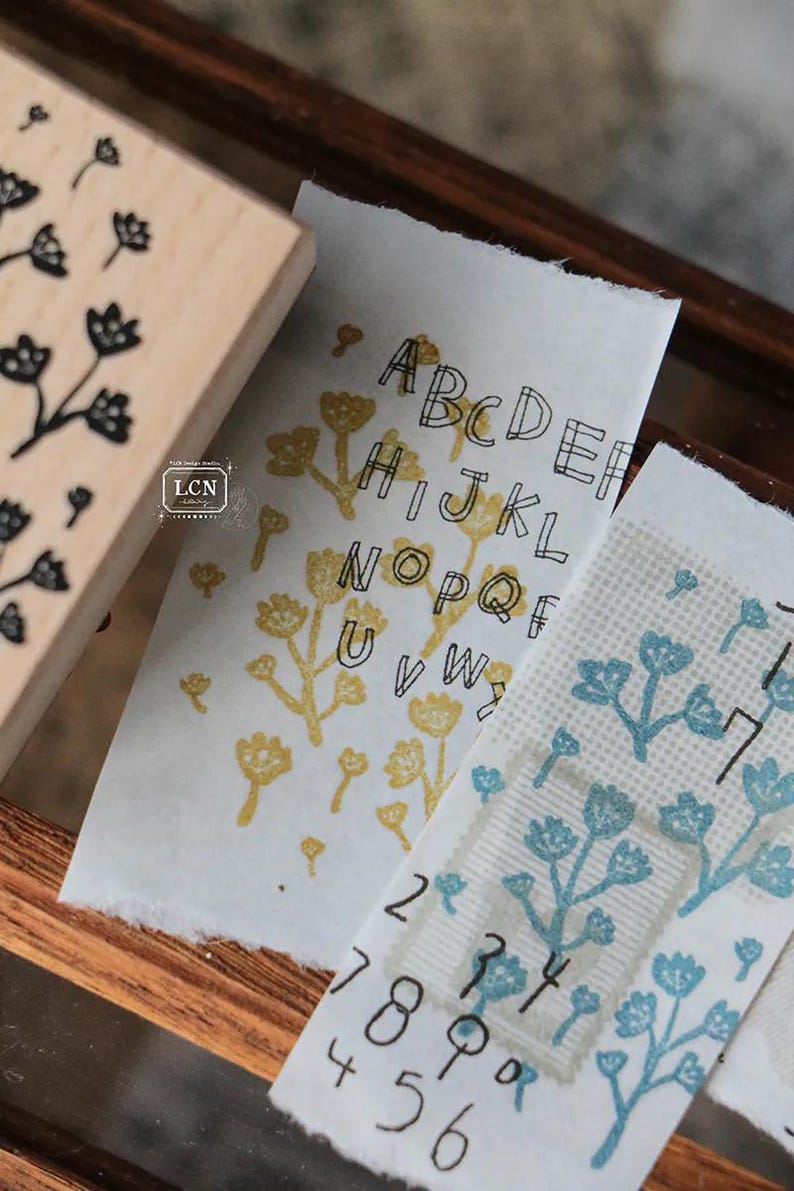 May include: Wooden floral stamp next to stamped paper with the alphabet and numbers. The paper has a torn edge and is stamped with yellow and blue floral designs. The stamp has a floral design.