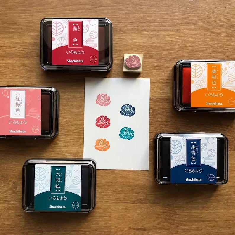 May include: A collection of Shachihata ink pads in various colors, including red, pink, orange, teal, and blue, with a rose stamp and stamped rose designs on a white card. The ink pads are in clear plastic cases.
