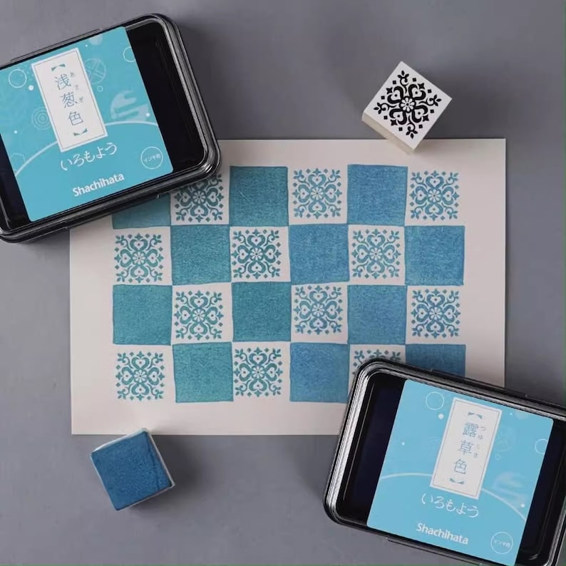 May include: A checkerboard pattern stamped in shades of blue on white paper. Two ink pads and a stamp are visible. The ink pads have the text "Shachihata" and Japanese text. The stamp has a black floral design.