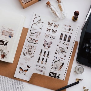 May include: An overhead shot of a workspace with stationery. A notebook, brown envelope, and a sheet of stickers with butterfly and botanical designs are visible. A vintage typewriter and small glass bottles with cork stoppers are also present.