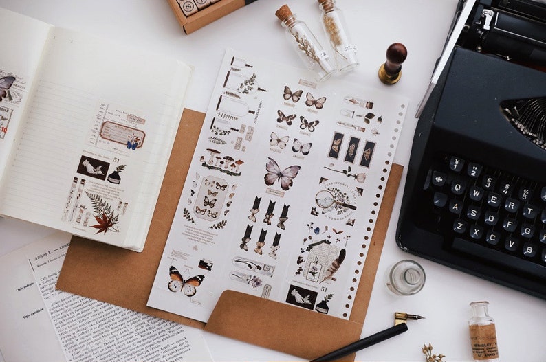 May include: An overhead shot of a workspace with stationery. A notebook, brown envelope, and a sheet of stickers with butterfly and botanical designs are visible. A vintage typewriter and small glass bottles with cork stoppers are also present.
