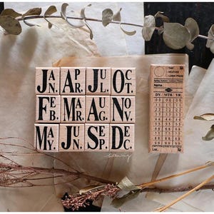 May include: Wooden stamps with the months of the year in a block format, alongside a smaller stamp with date and weather information. The stamps are arranged on a neutral-toned paper with dried foliage.
